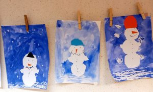 Kuvassa näkyy lasten tekemiä lumiukkoaskarteluja sinisellä pohjalla ja valkoisella kartongilla tehtynä.