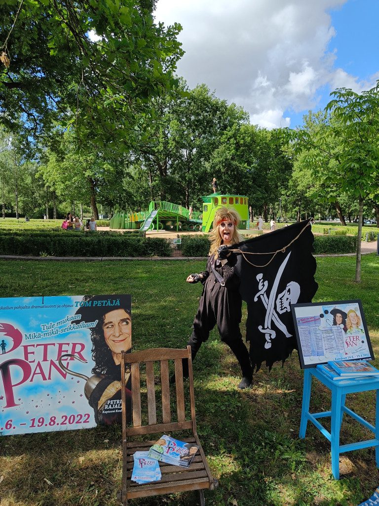 En person med piratdräkt håller en svart och vit piratflagga på axeln och räcker glatt ut tungan åt kameran. Framför personen står två stolar, en blå och en av trä. På stolarna ligger olika broschyrer som man har ställt framme. Ett parkområde och en lekplats syns i bakgrunden.