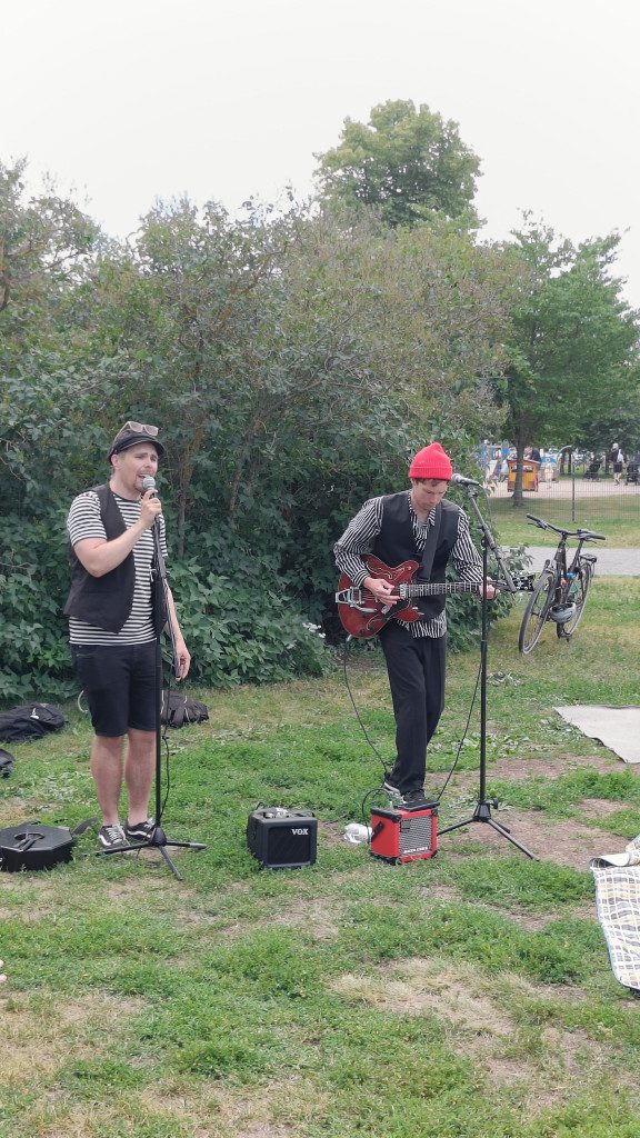 Två personer klädda i svart-vit-randiga skjortor och svarta västar uppträder framför en stor buske. Personen till vänster sjunger i mikrofonen medan personen till höger spelar gitarr. Den gitarrspelande personen har en röd mössa på huvudet.
