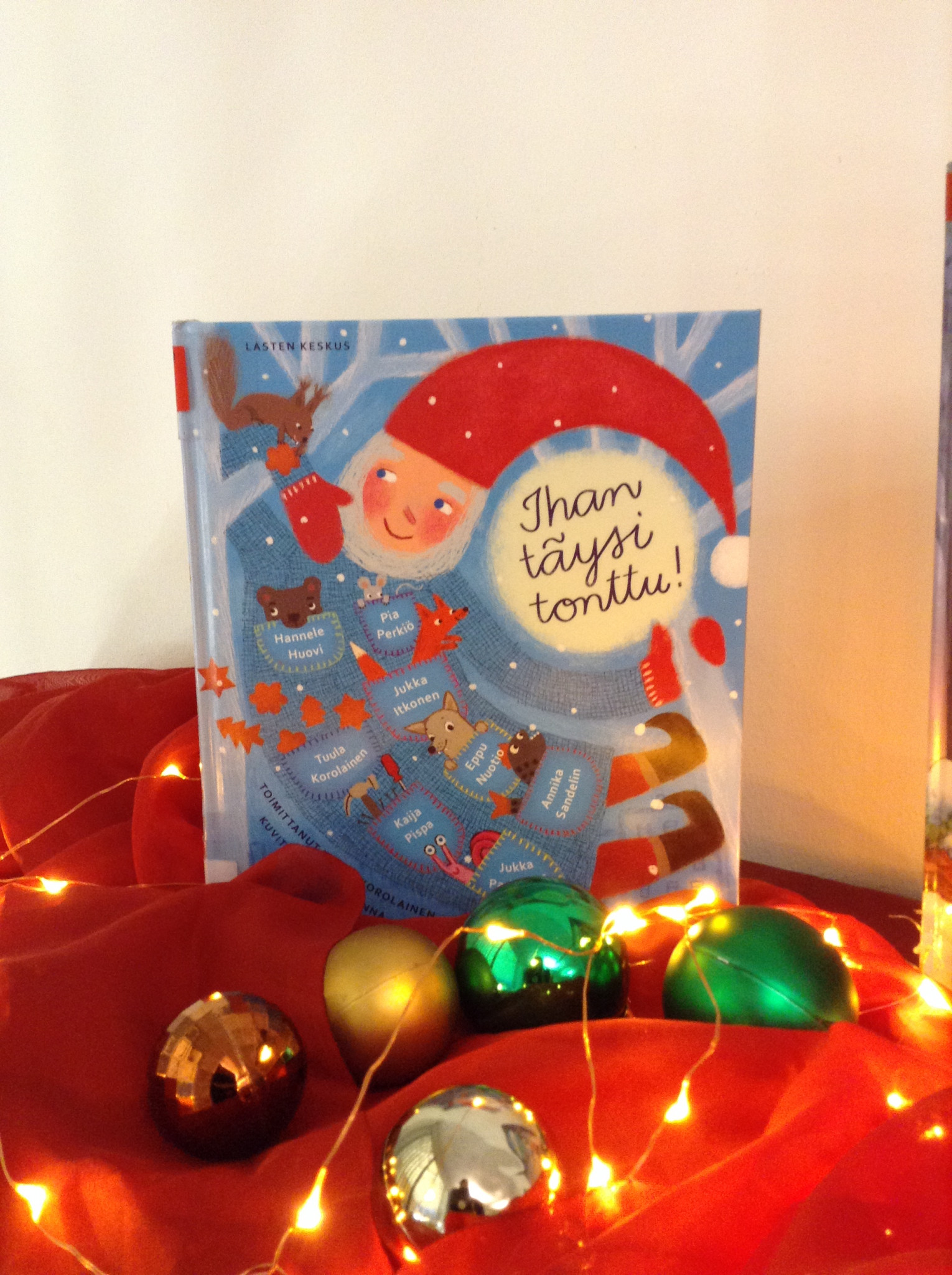 Jouluinen asetelma, jossa lasten joulukirja nojaa seinään punaisen kankaan päällä. Kirjan kannessa on iloisen näköinen tonttu. Kirjan edessä on joulupalloja ja jouluvalosarja.