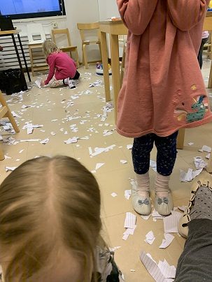 Kolme lasta kerää paperisilppua lattialta.
