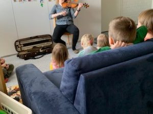 Aikuinen soittaa päiväpiirissä viulua ja lapset katsovat ja kuuntelevat etualalla.