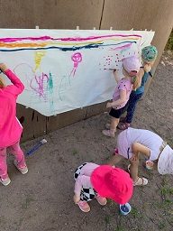 Iso paperi on ripustettu ulkona palloseinään ja siihen ryhmä lapsia maalaa vesiväreillä erilaisia kuvia.