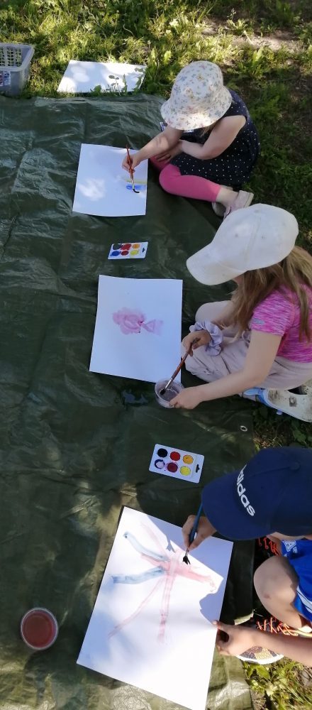 kolme lasta maalaamassa vesiväreillä ulkona pressun päällä