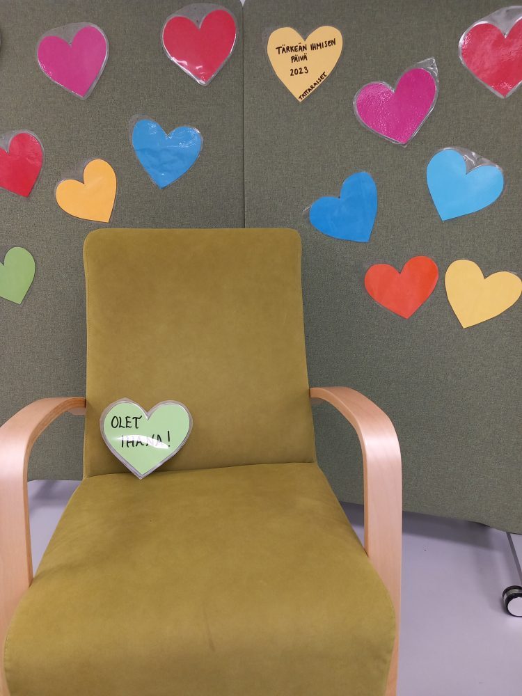 vihreä nojatuoli, taustalla vihreät sermit, joihin kiinnitetty värikkäitä sydämiä, tuolilla yksi sydän, johon kirjoitettu olet ihana