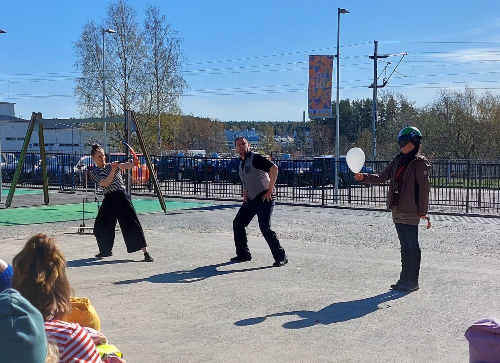 sirkustaiteilijat tähtäävät leikkijousipyssyllä ilmapalloon, joka on ihmisen kädessä