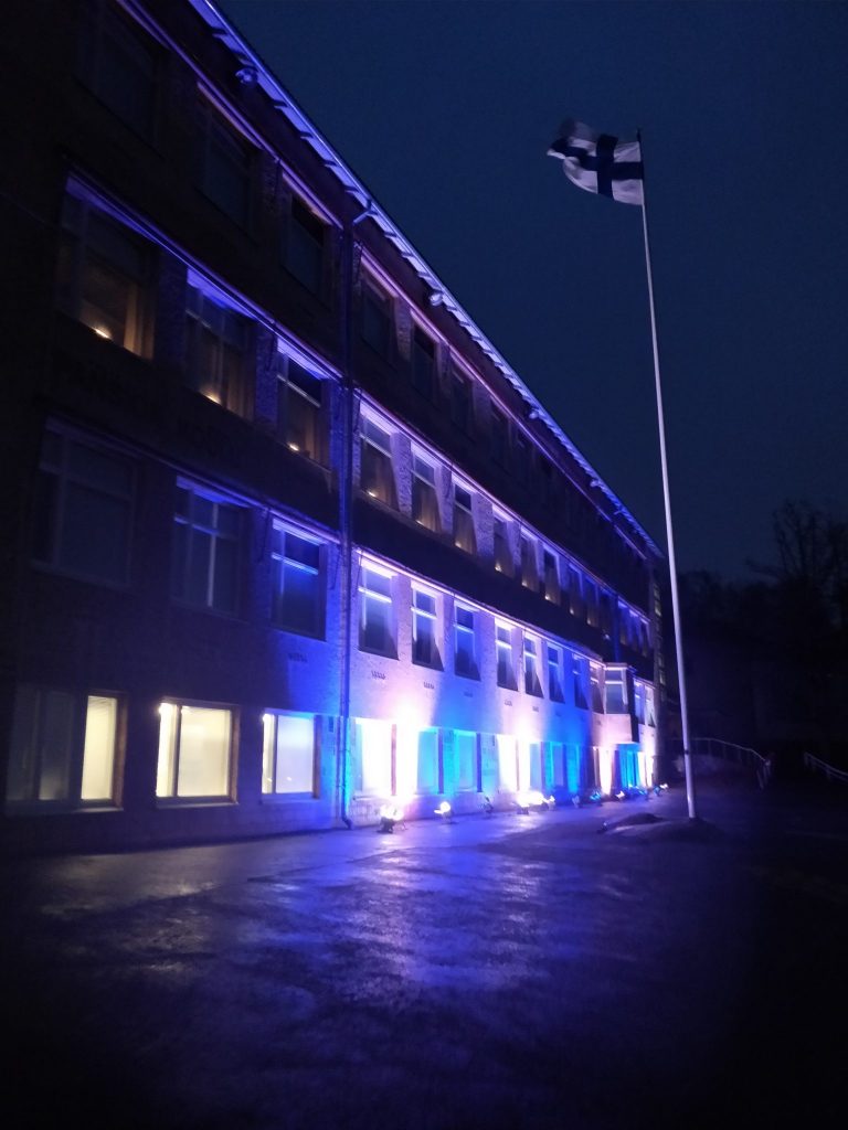 Suomen lippu liehuu lipputangossa koulun pihalla. Koulurakennus on valaistu.