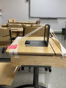 Kuvassa on pulpetti luokkahuoneessa. Pulpetin päälle on kiinnitetty maalarinteipillä iPad.