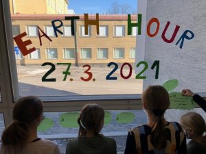 Ikkuna, jossa lukee Earth hour 27.3.2021. Ikkunassa on oppilaiden tekemiä papereita, joissa lukee oppilaiden antamia ilmastolupauksia. Kuvassa on muutamia lapsia kiinnittämässä lupauslappujaan.