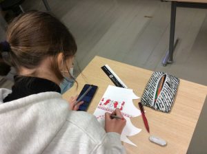 Kuvan vasemmassa reunassa on oppilas selin kameraan, jonka olan takaa näkyy pulpetti. Pulpetilla on valkoinen paperi, jolle oppilas piirtää puna-mustasäyistä Mezen-koristelua.