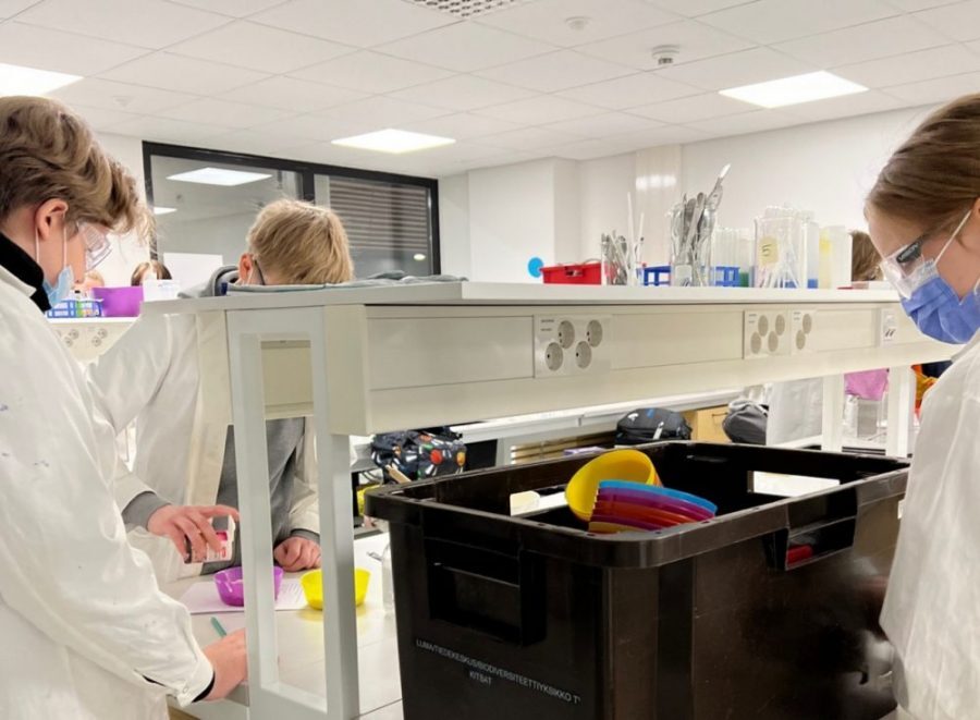 LUMA-keskuksessa. Oppilaita työskentelee laboratoriopöydän ääressä. Heillä on valkoiset suojatakit päällään.
