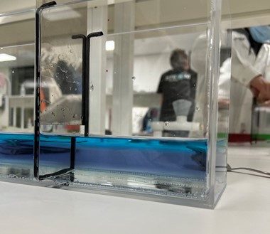 Kuvassa ollaan laboratoriossa. Pöydällä on iso lasinen laatikko, jossa on sinistä nestettä. Laatikon takana näkyy ihmisen ylävartaloa.