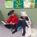 Kaksi opplasta istuu käytävän lattialla ja nojaa seinään. Toinen on pukeutunut merirosvon rooliasuun, toinen kissan viittaan. Molemmat ovat syventyneet lukemaan kirjaa.
