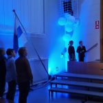 Sinivaloisesa hämärässä salissa seisoo Suomen lipun vieressä kolme oppilasta. Taaempana on valkoisia ilmapalloja ja viiden alla kaksi juontajaoppilasta.