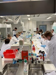 Oppilaita valkoisisa takeissaan molemmin puolin pitkää laboratorion tutkimuspöytää kokeita tekemässä.