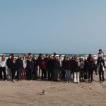 Nelisenkymmentä oppilasta seisoo harmaassa säässä tuulisella rantaviivalla. Takana vaahtopäinen meri ja edustalla hiekkarantaa.