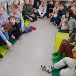 Paljon lapsia istuu puolipiirissä luokan lattialla vihreillä jumppamatoilla kuunnellen satua englanniksi.