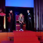 Punaisella lavalla seisoo 9-gaalan juontajiaPunaisella lavalla valtaistuimella istuu oppilaiden valitsema opettaja, vuoden kuningatar. Vieressä seisoo vuoden kuningas, miespuolinen opettaja.. Taustalla musta esirippu.