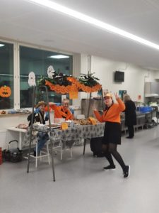 Kuvassa Halloween-juhlan buffetin pitäjiä koulun ruokasalissa. Kaksi tyttöä pukeutuneena Halloween-asuihin. Myyntipisteen edessä aikuinen Halloween-asussa.