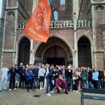 Ryhmä oppilaita seisoo kirkon edustalla. Näkyvissä suuri oranssi lippu, jossa lukee Nieuwe Kerk.