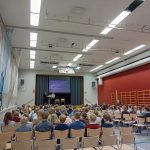 Oppilaita takaapäin kuvattuna alasalissa. Salin lavalla kaksi henkilöä kertomassa runoudestaan.