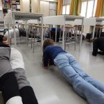 Lattialla, luokan pöytien alla oppilaita etunojassa, lankutusasennossa.