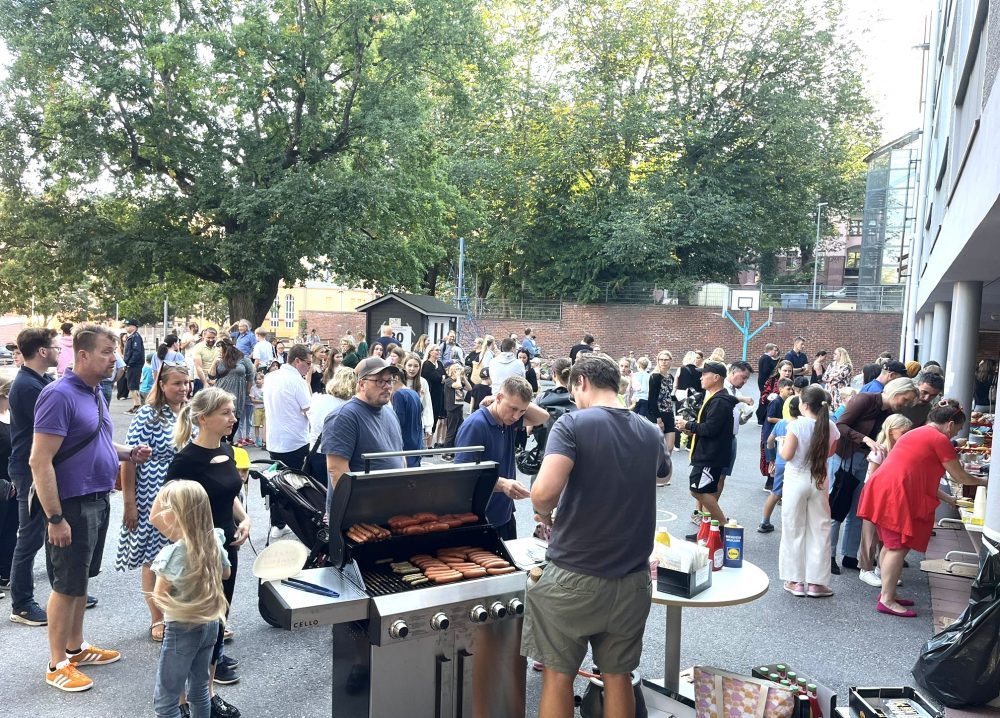 Etualalla mies grillaa makkaraa. Takana aikuisia ja lapsia koulun pihalla.