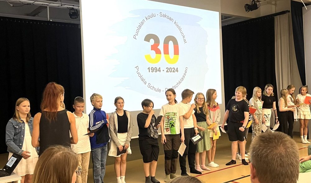 Oppilaita rivissä juhlasalin valkokankaan edessä. Valkokankaalla on saksan kieliluokkien 30-vuotislogo.