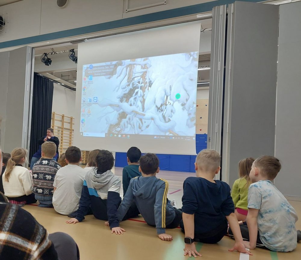 Lapset istuvat liikuntasalissa katsomassa isoa näyttöä, jossa on talviaiheinen kuva.