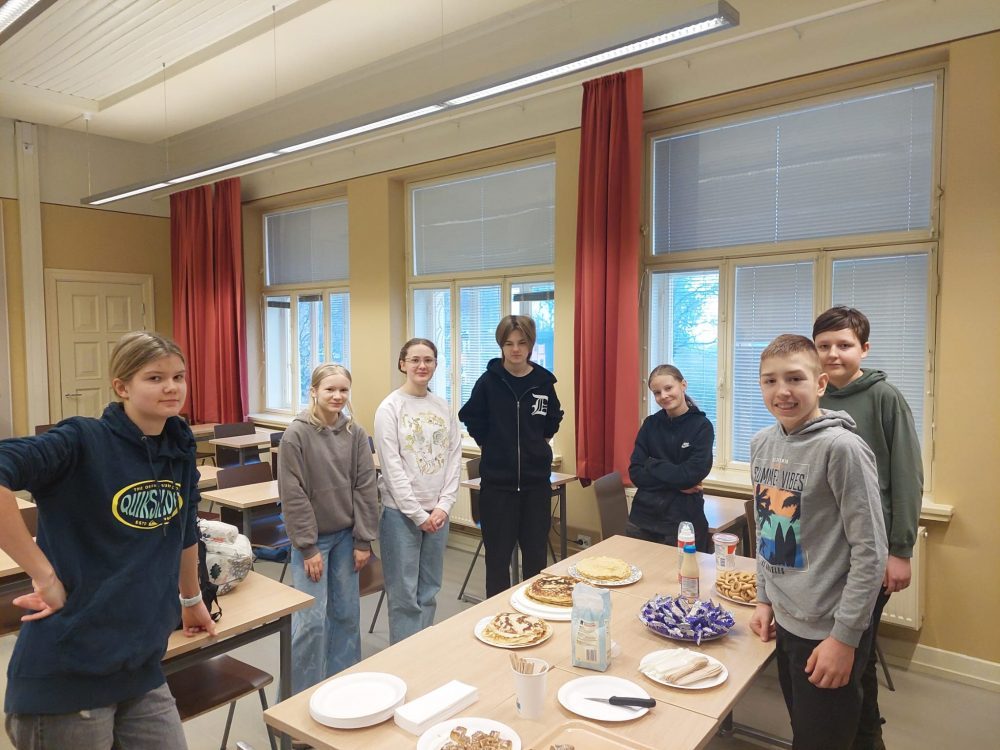Oppilaat seisovat ison pöydän ympärillä. Pöydällä on blinejä, karkkeja ja muita herkkuja.