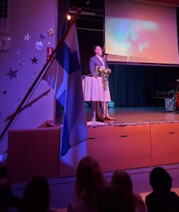 Kuvassa seisoo lavalla puhujanpöydän oikealla puolella juhlapuhettä pitävä mies. Etualalla vasemmalla kuva-alassa liehuu Suomen lippu. Taustalla näkyy valkokankaalle heijastettua kuvaa epäselvästi.