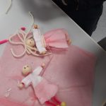 Kuvassa vaalenapunaista tarlataania, jonka päällä kaksi piipunrassista ja kangaspaloista askarreltua ihmishahmoa, joilla on isompi puuhelmi päänä. Käsinä ja jalkoina on pienet puuhelmet.