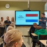 Keskellä kuvaa on näyttö, jossa näkyy Hollannin lippu. Näytön vasemmalla puolella on 3 oppimasta kertomasa etualalla olevalle luokalle.