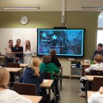 Vasemmalla puolella kuvaa kolme oppilasta selittää jotakin etualan luokalle. Keskellä näkyy Powerpoint-dia