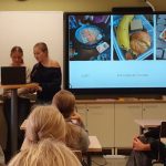 Vasemmalla puolella kuvaa kolme oppilasta selittää jotakin etualan luokalle. Keskellä näkyy Powerpoint-dia, jossa on ruokakuvia.