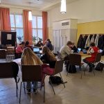 Kuvassa näkyy koulun aula, jossa useita pöytiä, Niiden ympärillä on oppilasryhmiä tietokoneiden kanssa.