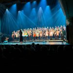 Alakoulun musiikkiluokkien kuorot lavalla. Lapsilla värikkäät vaatteet. edessä mustaa pimeyttä. Yläosassa kuvaa voimakkaat sinuvalokaartiot ylhäältä alas.