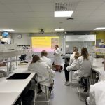 Oppilaita kuuntelemassa opettajan esitystä. Opilaat nojailevat valkoiset laboratoriotakit yllään pöytiin.