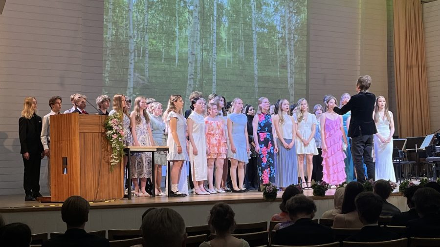 Noin 30 oppilaan kuoro konserttitalon lavalla iloisissa kesämekoissaan laulamassa ja kuoronjohtaja edustalla johtamassa kuoroa.