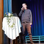 Puheen pitäjä pyöreän valkoisen liinan verhoaman coctailpöydn ääressä. Pöydällä sinivalkoinen kukka-asetelma.
