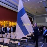Keskellä kuvaa Suomen lippu jota kannattele partiopukuun pukeutunut oppilas. Hänen takanaan kaksi airuetta.