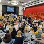 Oppilaita istumassa liikuntasalin lattialla takaapäin kuvattuna. Edessä näyttämöllä ohjelmansuorittajia.