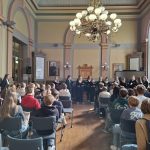 Kuoro laulaa juhlasalin etuosassa. Kuva otettu salin takaa, joten edustalla istuu oppilaita kuuntelemassa selin.