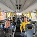Kuva on bussin sisältä, jossa etu alalla on opettajia ja takana istumassa on oppilaita. Porukka on iloinen ja jännittynyt.