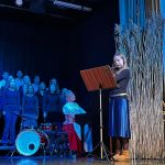 Taustalla kuoro, keskellä pianoa soittava kansallispukuinen opettaja ja oikealla huilua soittava tyttö.